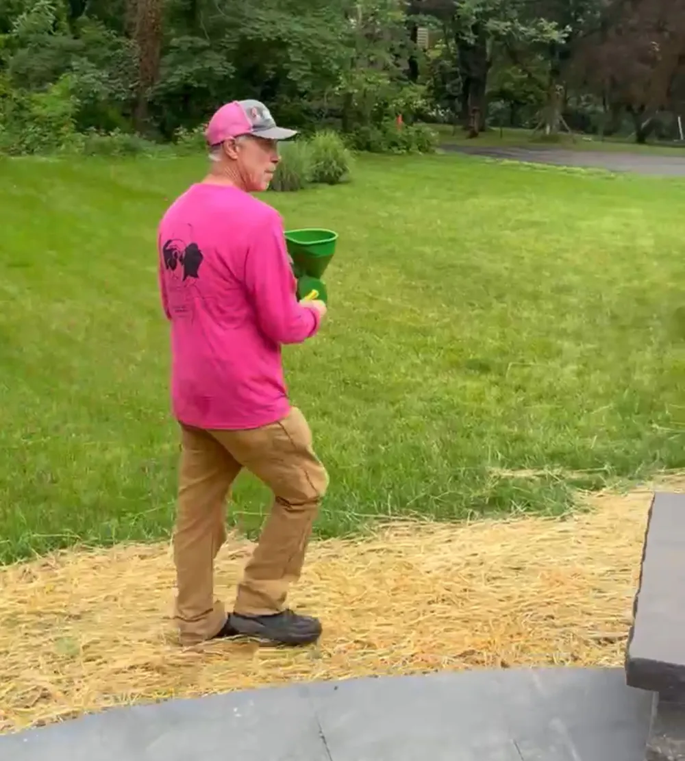 Full Circle Services technician in a pink shirt applying granular lawn fertilizer with a hand spreader