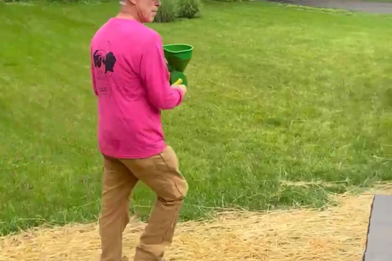 Full Circle Services technician in a pink shirt applying granular lawn fertilizer with a hand spreader